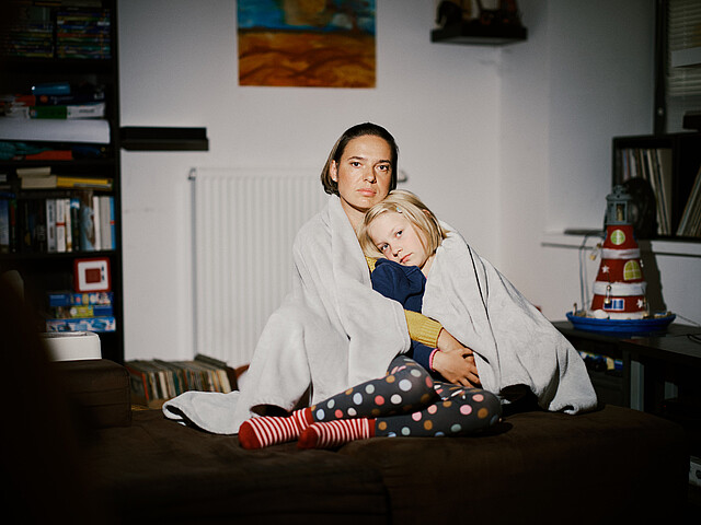 Eine Mutter sitzt mit ihrer Tochter in eine Decke gehüllt auf einer Couch. Im Hintergrund ist ein Wohnraum zu sehen.