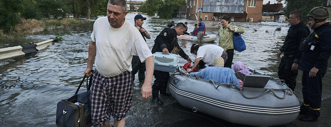Ein Mann trägt eine Reisetasche, er kommt der Kamera entgegen. Im Hintergrund ist alles überschwemmt. Menschen werden in Schlauchbooten evakuiert.