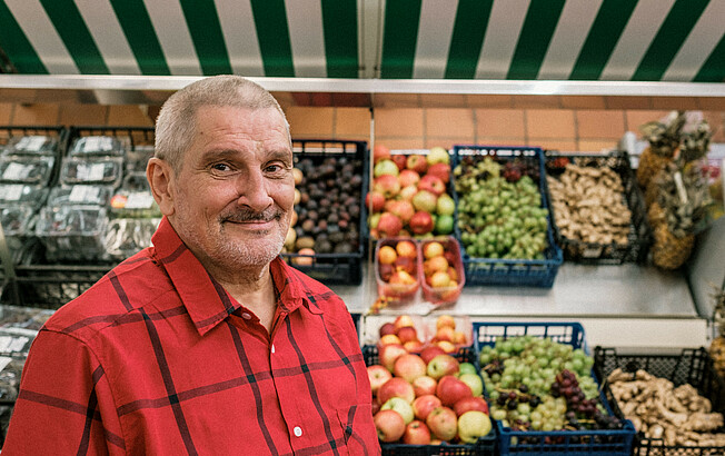 Mann blickt zur Kamera, im Hintergrund ein Obst- und Gemüseregal