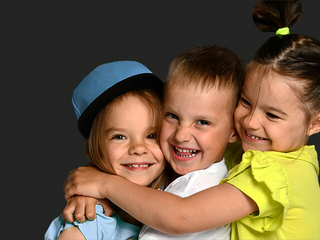 Drei Kinder umarmen sich und lachen in die Kamera, der Hintergrund ist schwarz.