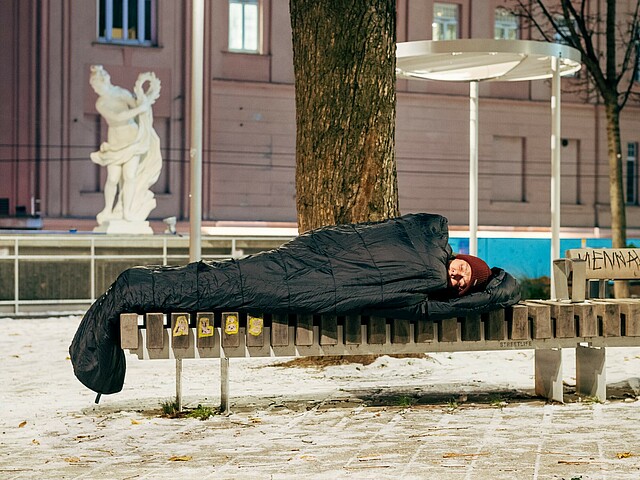 Eine Person schläft auf einer Parkbank, sie ist in einen Schlafsack gewickelt. Es ist winterlich.