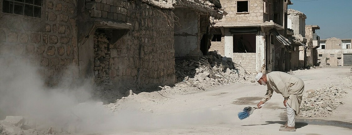 Es sind Ruinen von Häusern zu sehen, zerstört von den Erdbeben. Ein Mann steht gebückt auf der Straße und kehrt. 