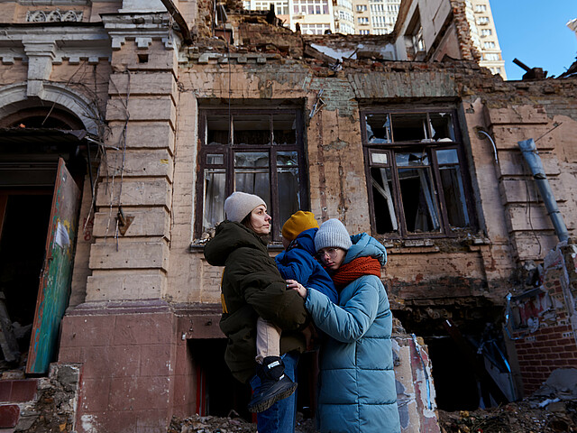Eine Mutter hält ein Kleinkind am Arm, daneben steht noch eine Jugendliche, sie umarmt die beiden. Sie tragen Winterjacken und Hauben. Im Hintergrund ist ein vom Krieg zerstörtes Haus zu erkennen.