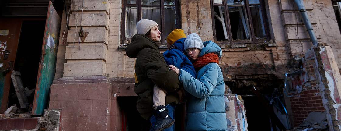 Eine Mutter hält ein Kleinkind am Arm, daneben steht noch eine Jugendliche, sie umarmt die beiden. Sie tragen Winterjacken und Hauben. Im Hintergrund ist ein vom Krieg zerstörtes Haus zu erkennen.
