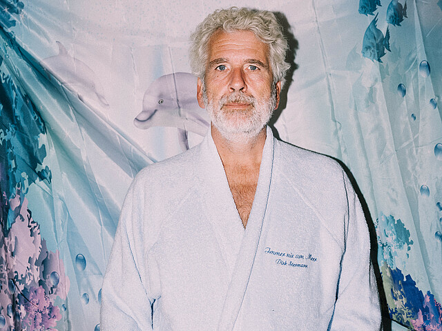 Dirk Stermann steht im Bademantel vor einem Duschvorhang