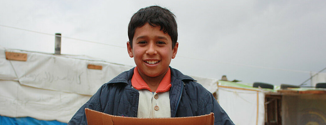 Ein Flüchtlingskind befindet sich im Libanon in einem Zeltlager. Er hält eine große Schachtel in den Händen. 
