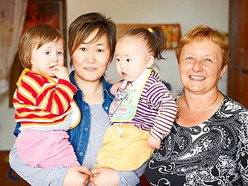 Zwei Frauen, die beide einen kurzhaarschnitt tragen, und zwei Säuglinge sind zu sehen. Dabei hat die jüngere Frau die zwei Babies auf dem Arm. Bekleidet sind alle mit bunten Farben. 
