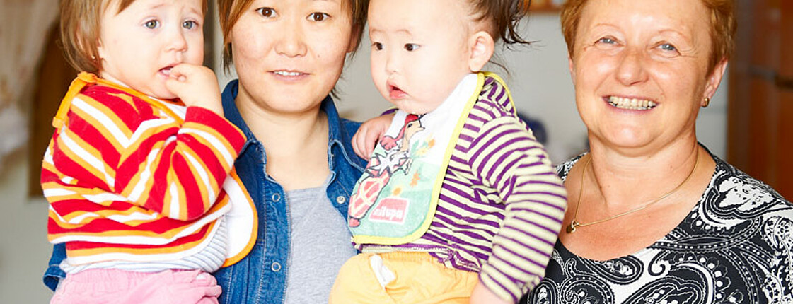 Zwei Frauen, die beide einen kurzhaarschnitt tragen, und zwei Säuglinge sind zu sehen. Dabei hat die jüngere Frau die zwei Babies auf dem Arm. Bekleidet sind alle mit bunten Farben. 