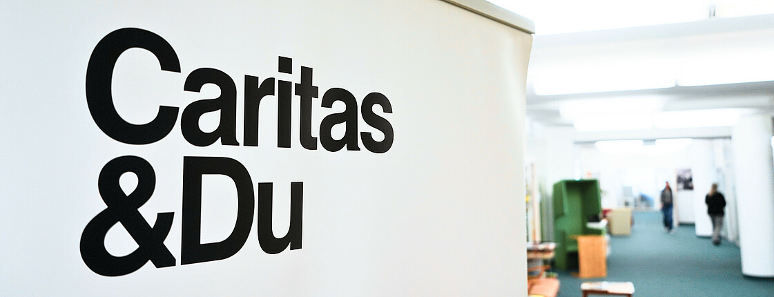 Schriftzug "Caritas & Du" auf Flipchart, im Hintergrund ist verschwommen ein Büro zu erkennen
