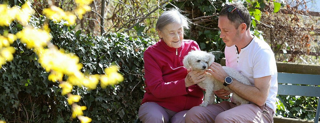 Ältere Dame und Freiwilliger sitzen auf einer Parkbank. Sie haben einen kleinen weißen Hund am Schoß, den sie beide streicheln.