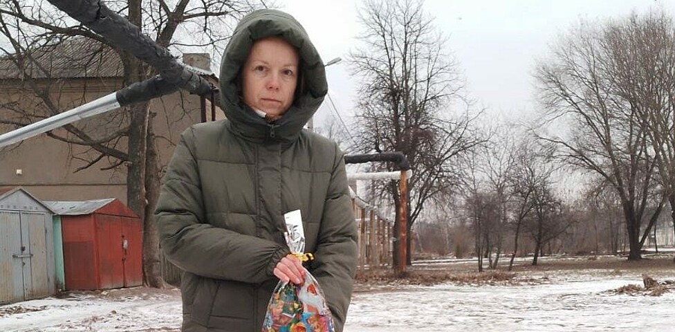 Eine Person in winterlicher Kleidung steht mit zwei Plastiktüten vor sich draußen. Die Landschaft ist schneebedeckt