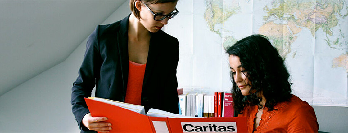 Zwei junge, erwachsene Frauen sind in einem Büro. Beide sind in rot gekleidet. Eine zeigt der anderen einen roten Ordner, auf dem das CARITAS Logo zu sehen ist. Eine der beiden Frauen trägt eine Brille.