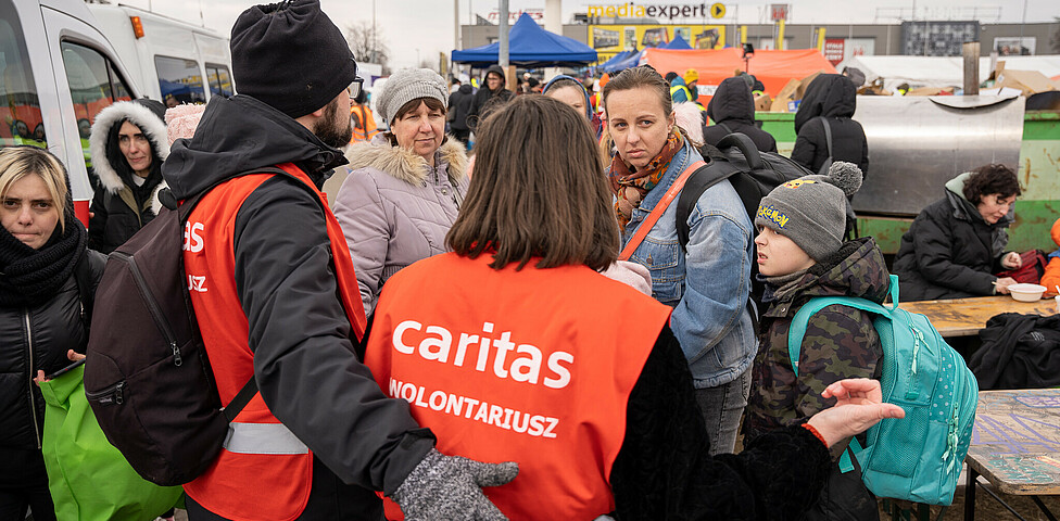Man sieht zwei Freiwillige der Caritas von hinten, sie tragen rote Westen mit Caritas-Logo, sie sprechen mit ankommenden Geflüchteten aus der Ukraine